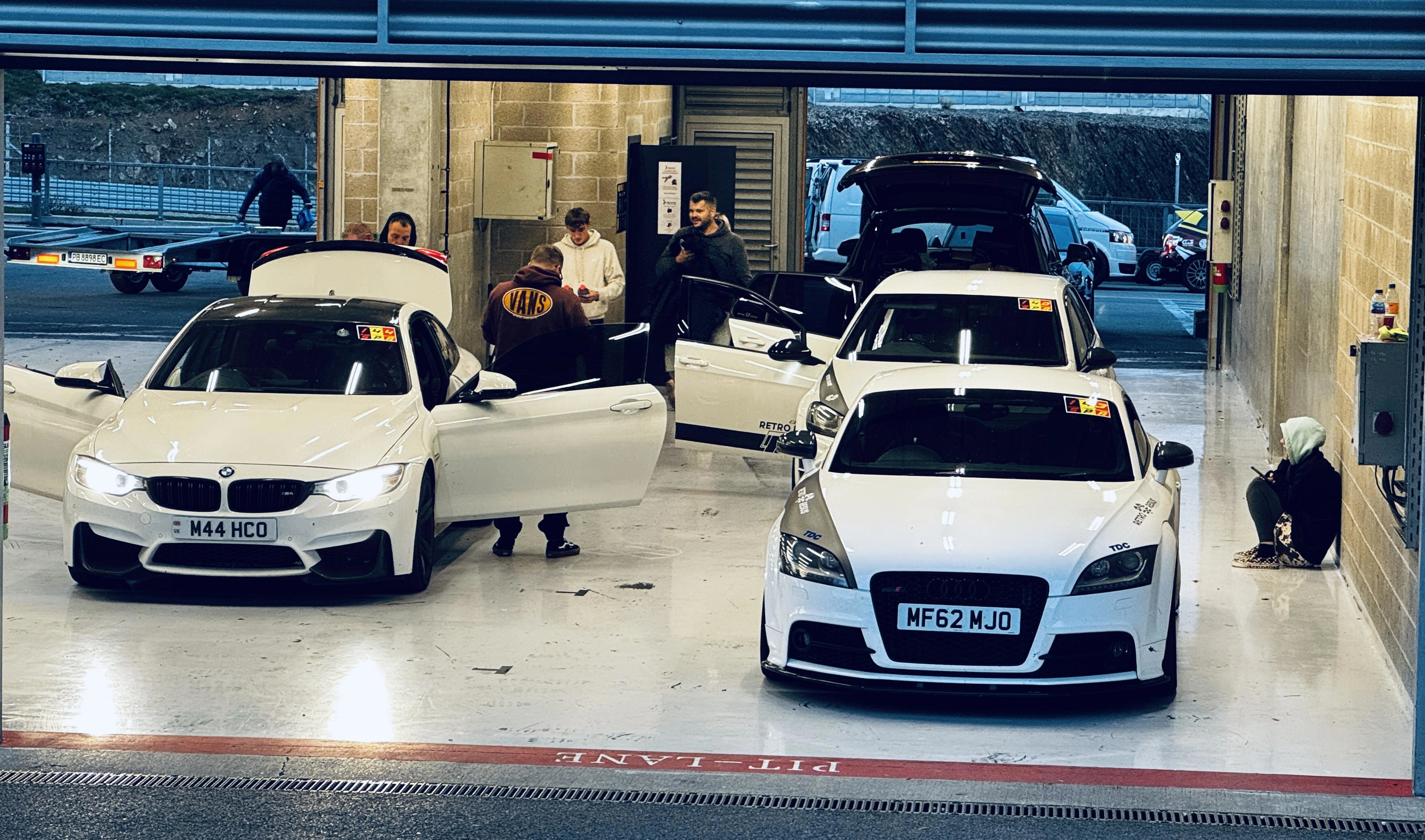 Garage with 4 cars with their doors open, there are peope gathered around them